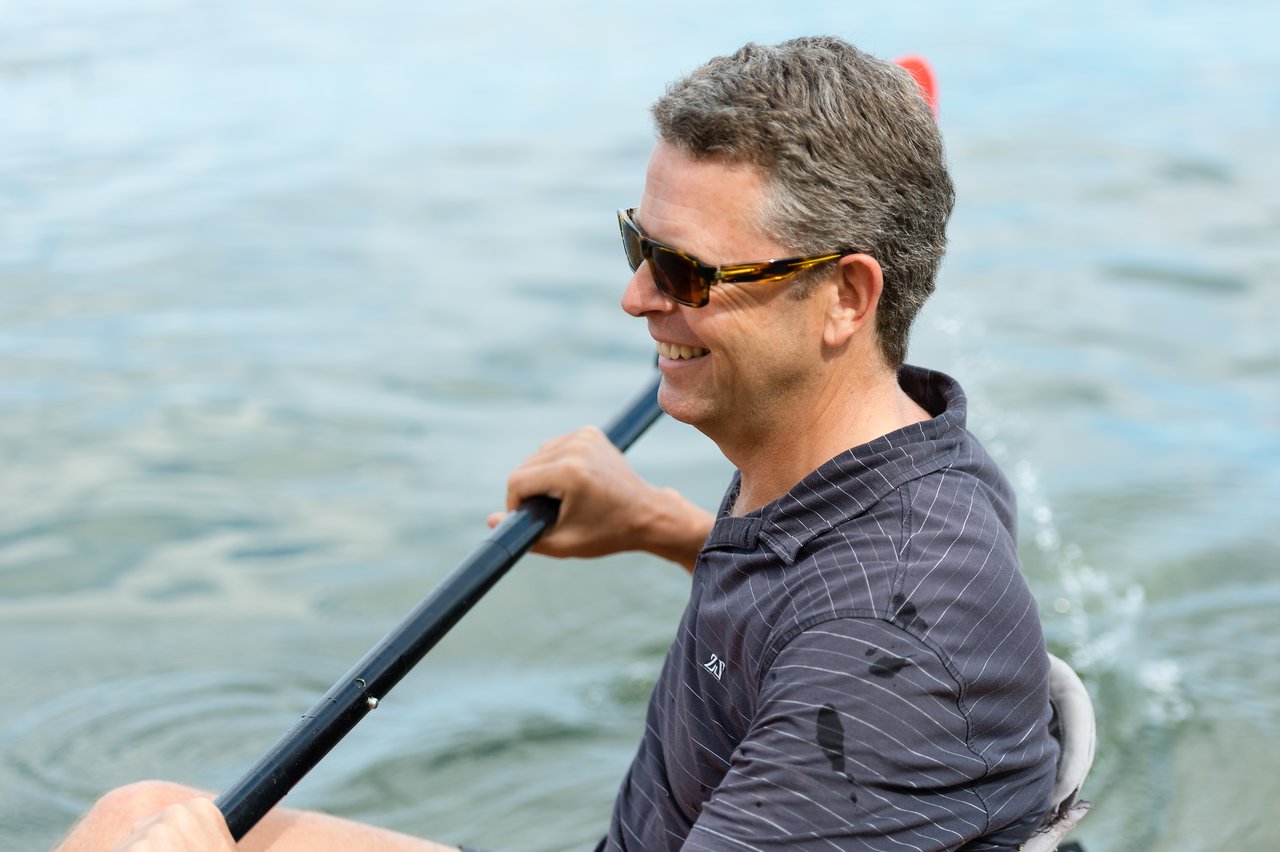 A man wearing sunglasses smiles while paddling a kayak on the water.