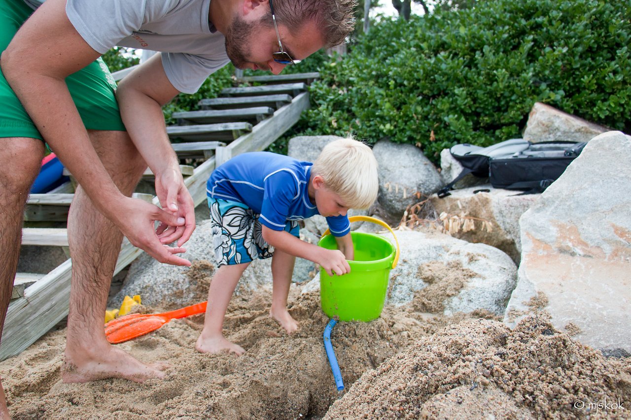 A young child in a blue shirt scoops sand into a green bucket while an adult watches nearby.