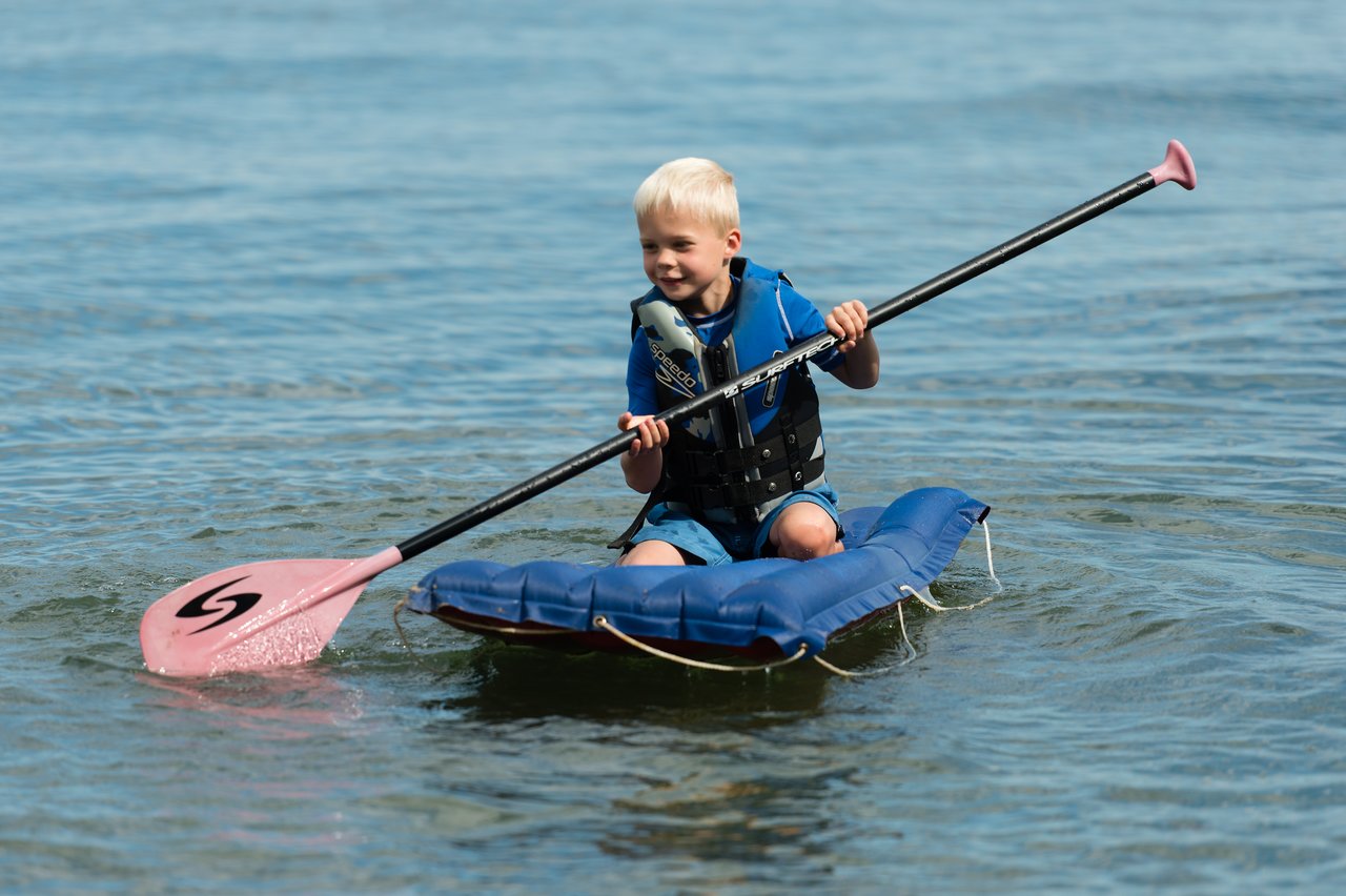 A young child wearing a life jacket paddles on a small inflatable raft in shallow water.