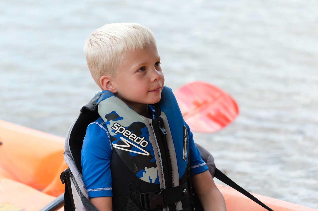 A young child wearing a life jacket sits in a kayak near the water, with a paddle in the background.
