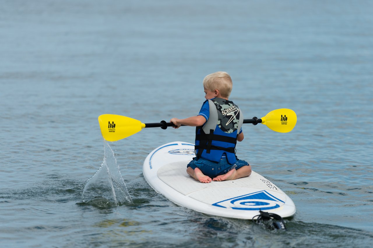 A young child wearing a life jacket paddles on a surfboard in calm water.