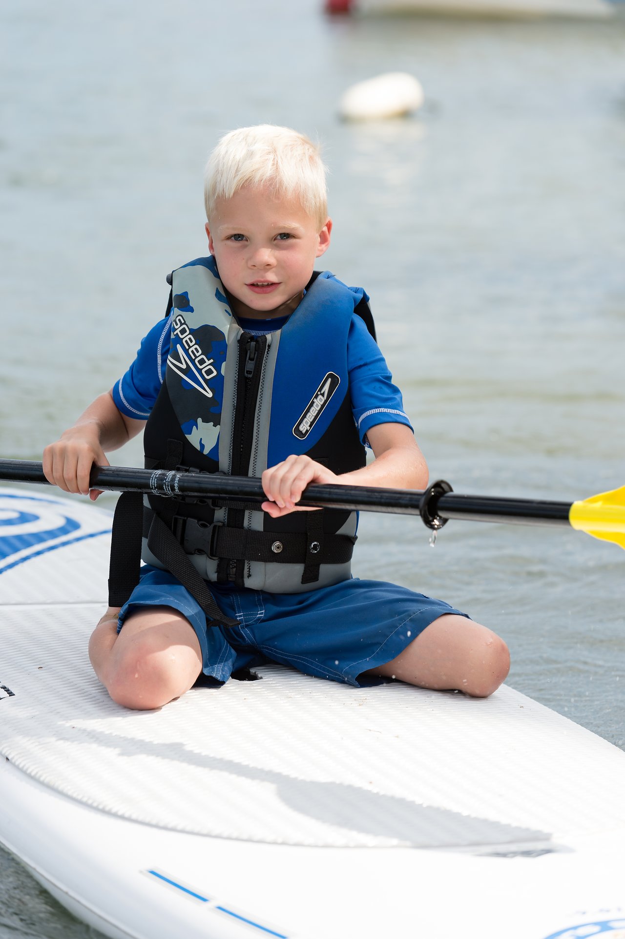 A young child wearing a life jacket sits on a paddleboard, holding a paddle over the water.