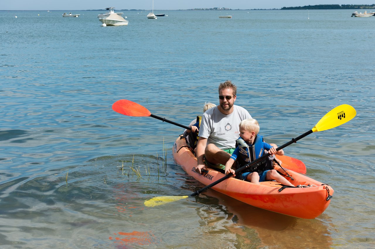 A man and two children paddle a bright orange kayak in shallow water near the shore.
