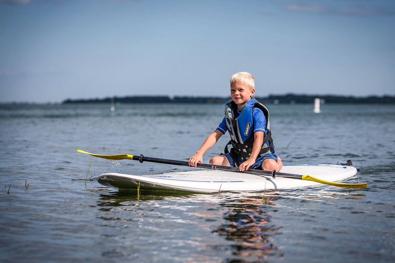 A young child wearing a life jacket sits on a paddleboard in shallow water, holding a paddle.
