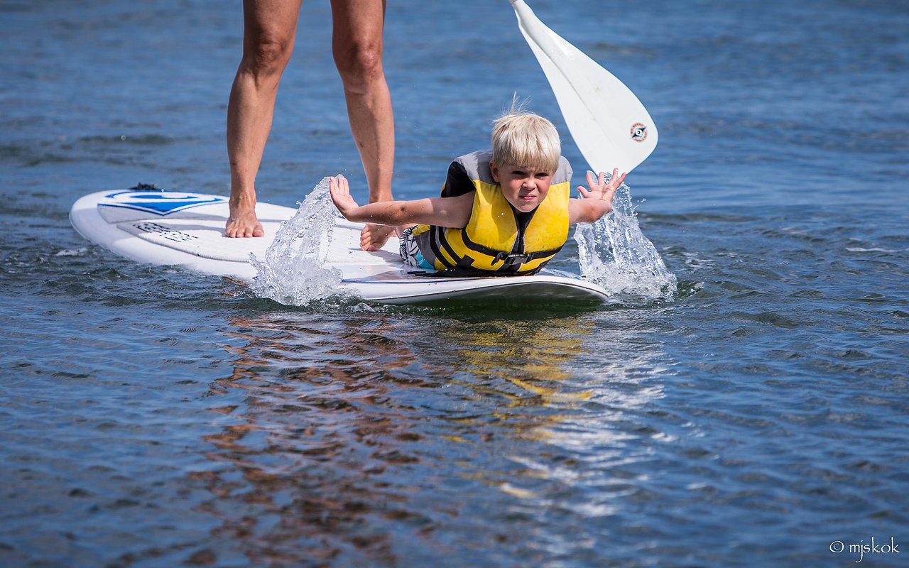 A child in a life jacket lies on a paddleboard, splashing water, while an adult stands behind them.