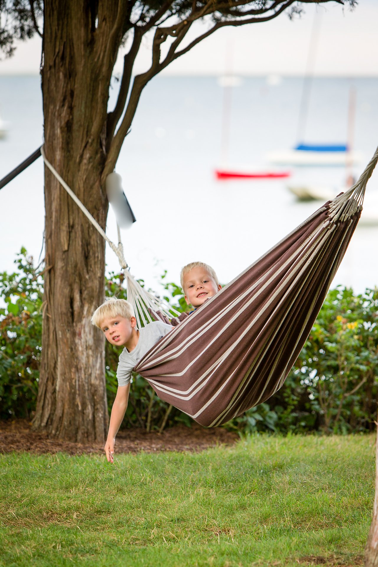Two young boys lie in a striped hammock, one reaching toward the grass, with boats visible in the background.