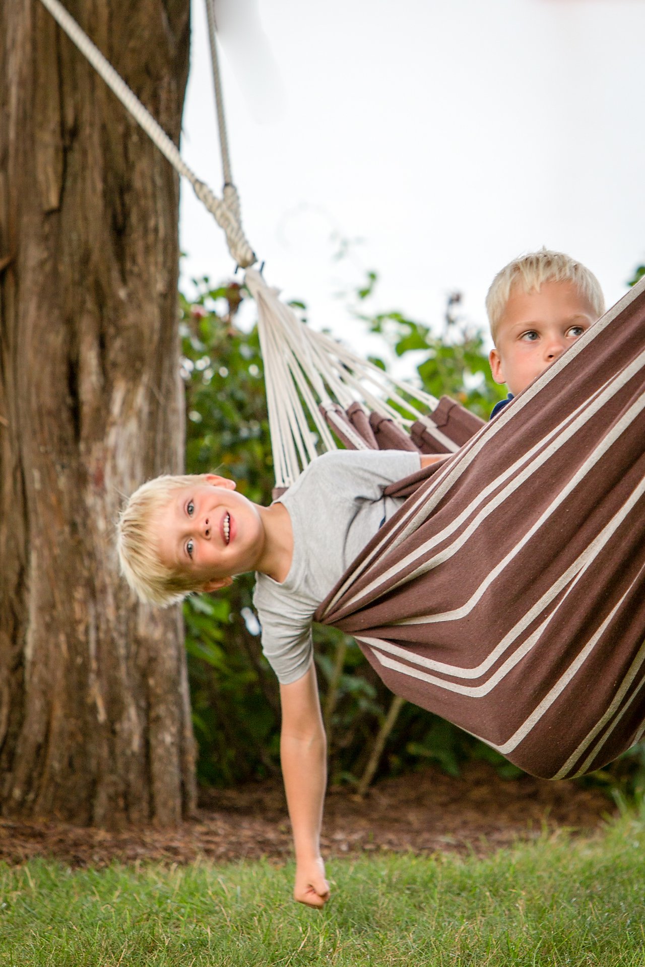 Two young boys sit in a striped hammock, one leaning out and smiling while the other looks to the side.