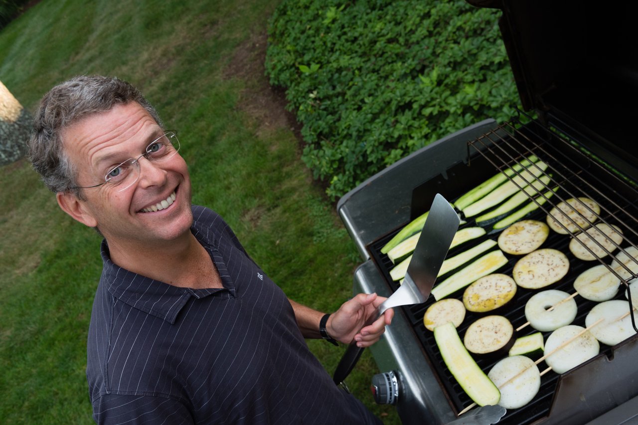 A man smiles while grilling sliced vegetables on a barbecue in a grassy backyard.