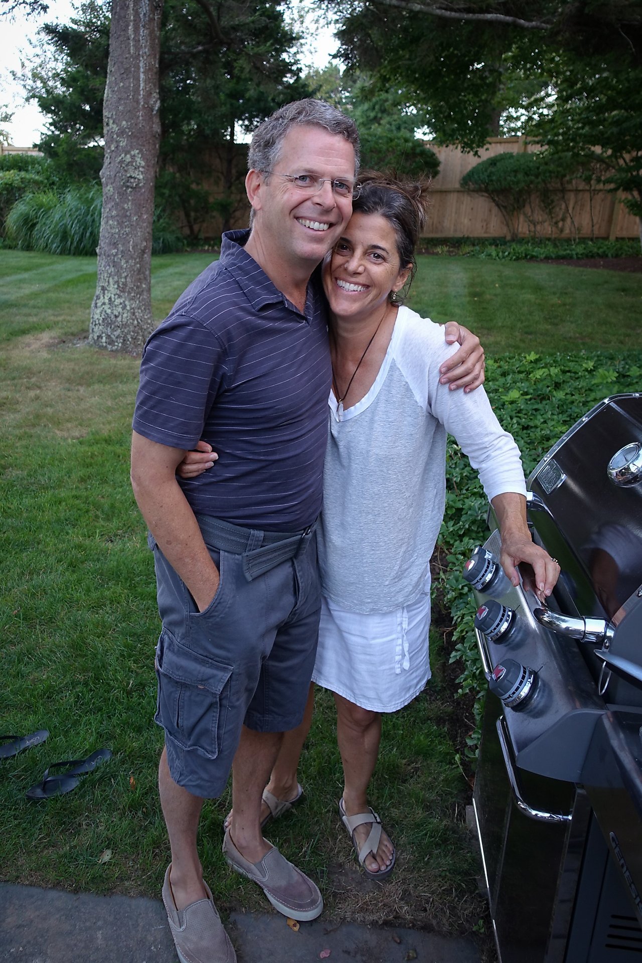 A smiling couple stands together by a grill in a backyard, with one person adjusting the grill knobs.