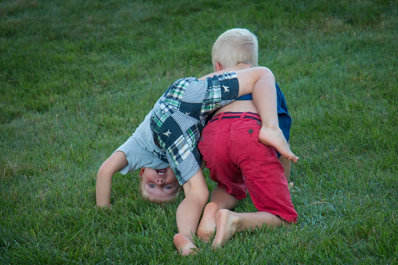 Two young children play on the grass, one upside down and smiling while the other leans forward.