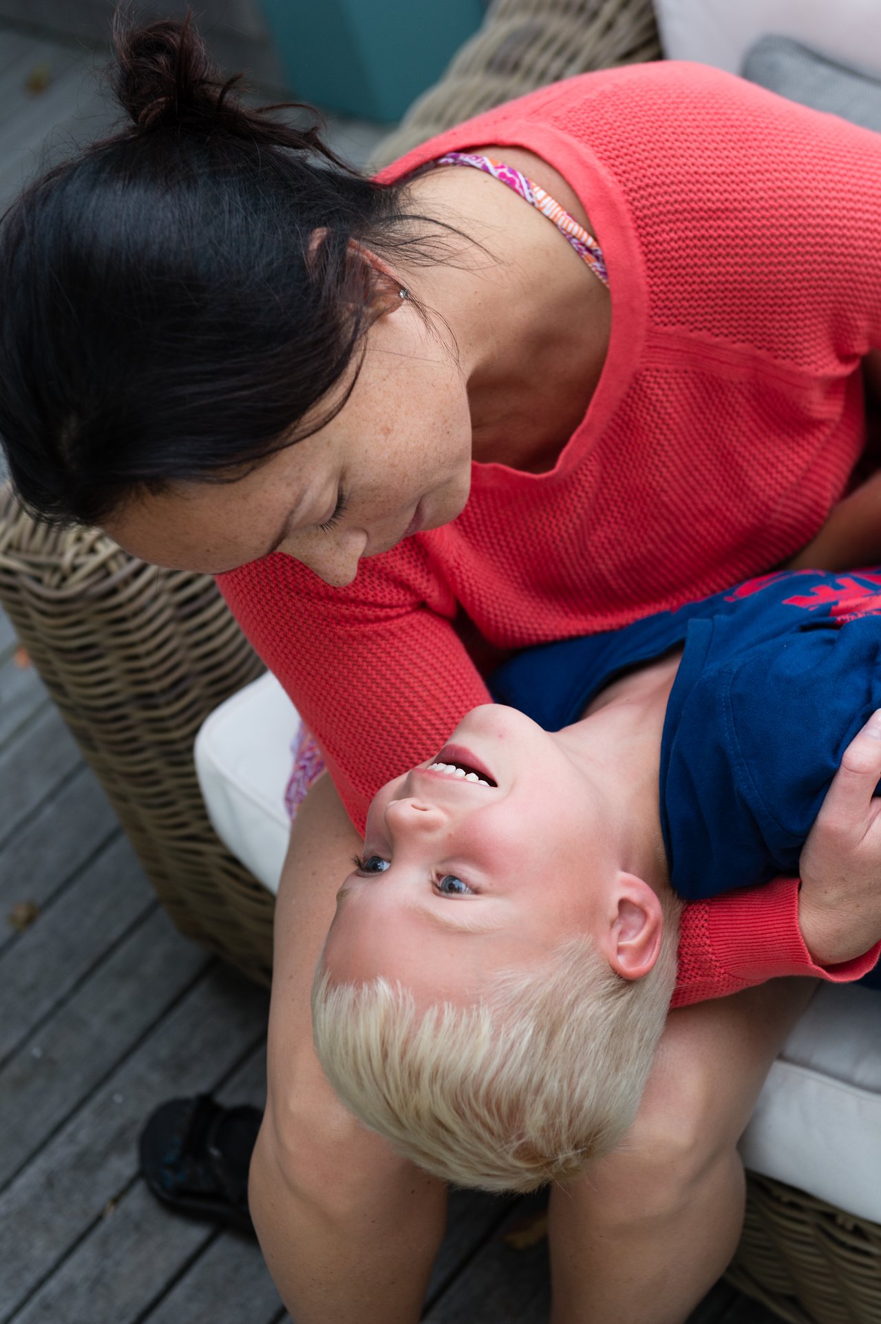 A woman in a red sweater holds a smiling child upside down on her lap while sitting on a wicker chair.