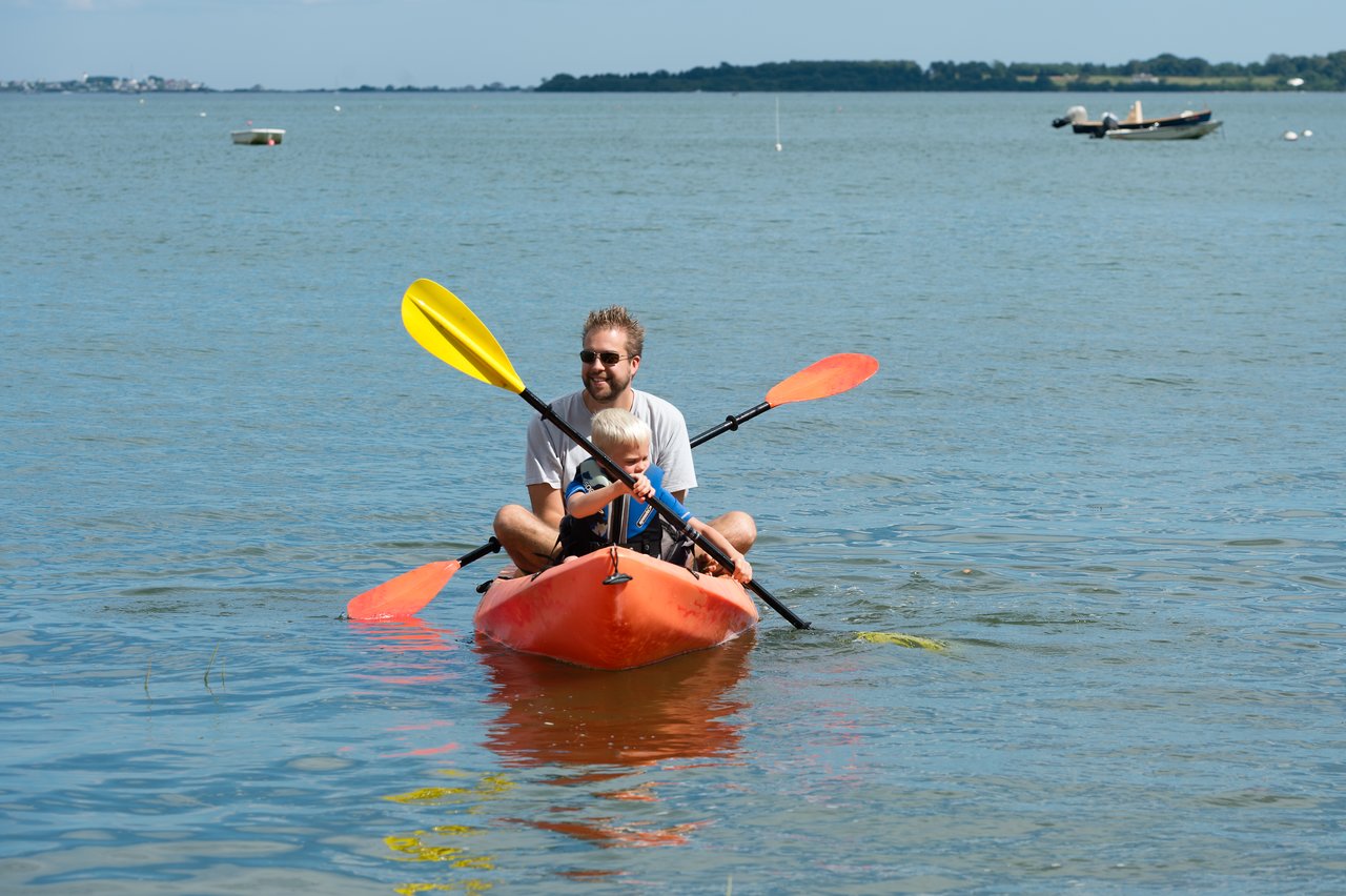 A man and a child paddle a red kayak in calm water on a sunny day.