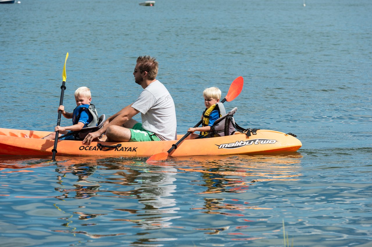 A man and two young children paddle an orange kayak on calm water, all wearing life jackets.