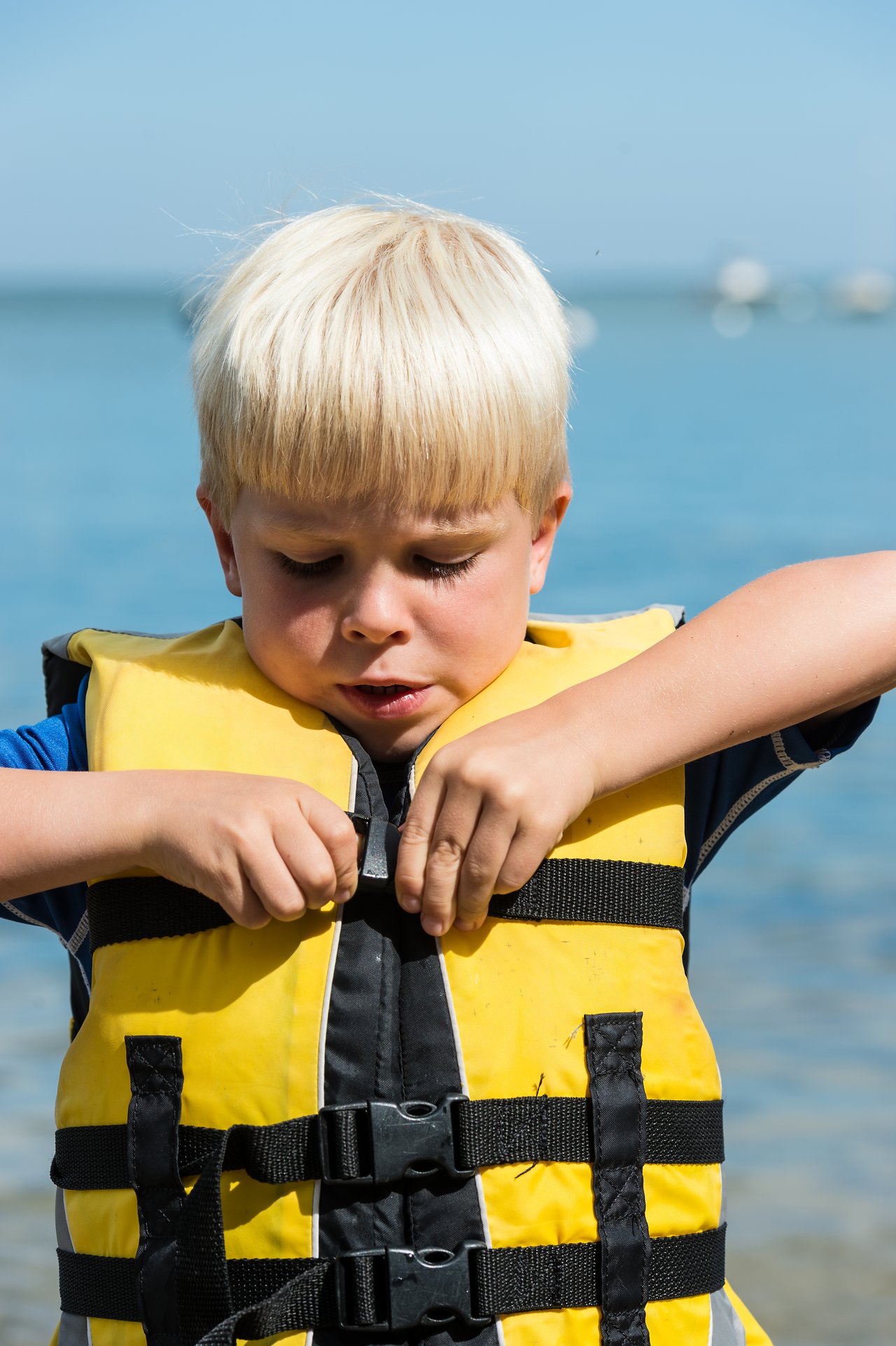 A young child in a yellow life jacket fastens the buckle while standing near the water.