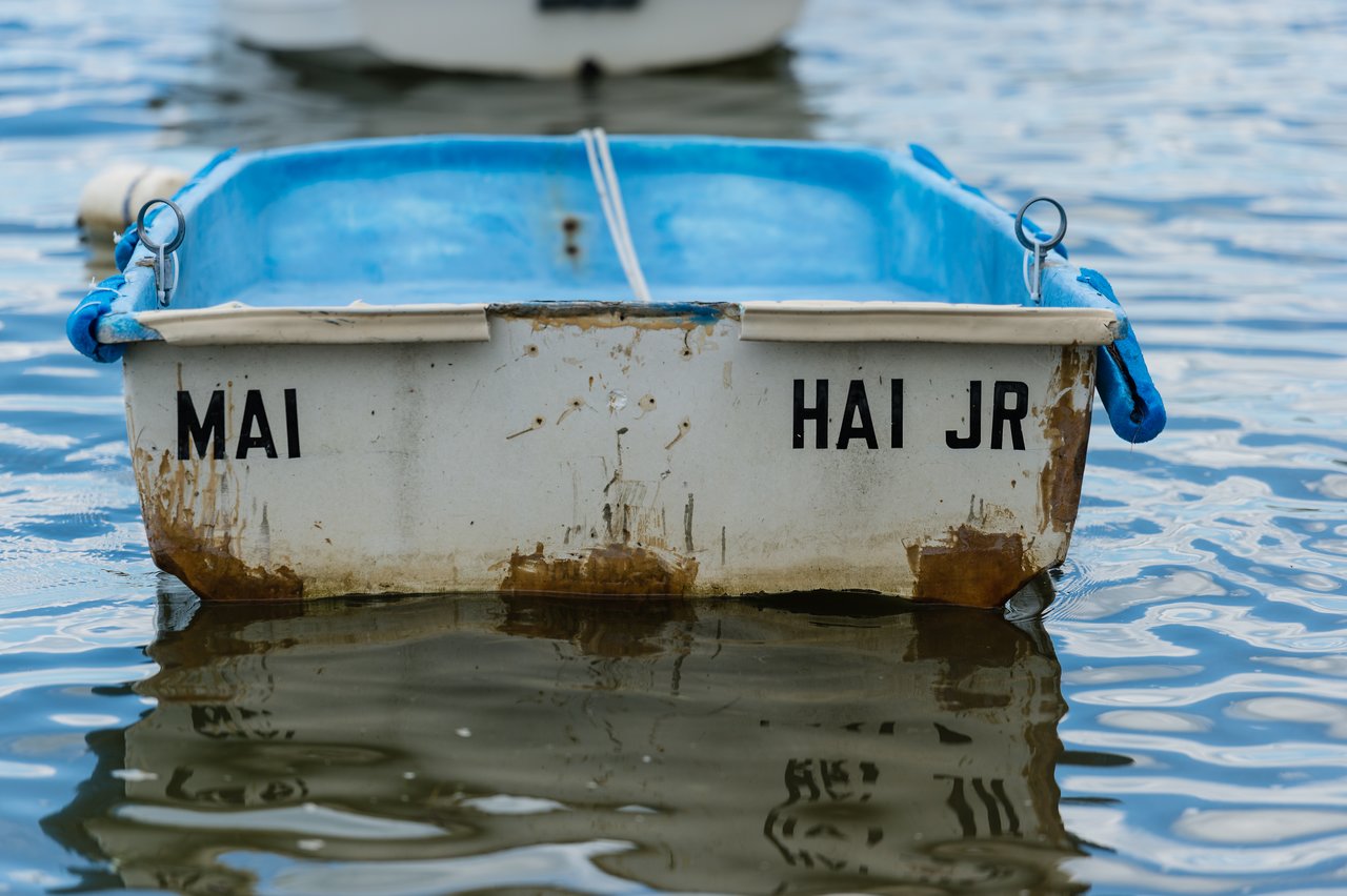 A small, weathered boat with "MAI HAI JR" on the back floats on calm water.