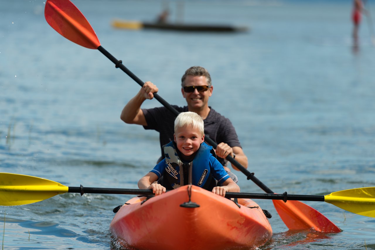 A man and a child paddle a bright orange kayak together on calm water, both wearing life jackets.