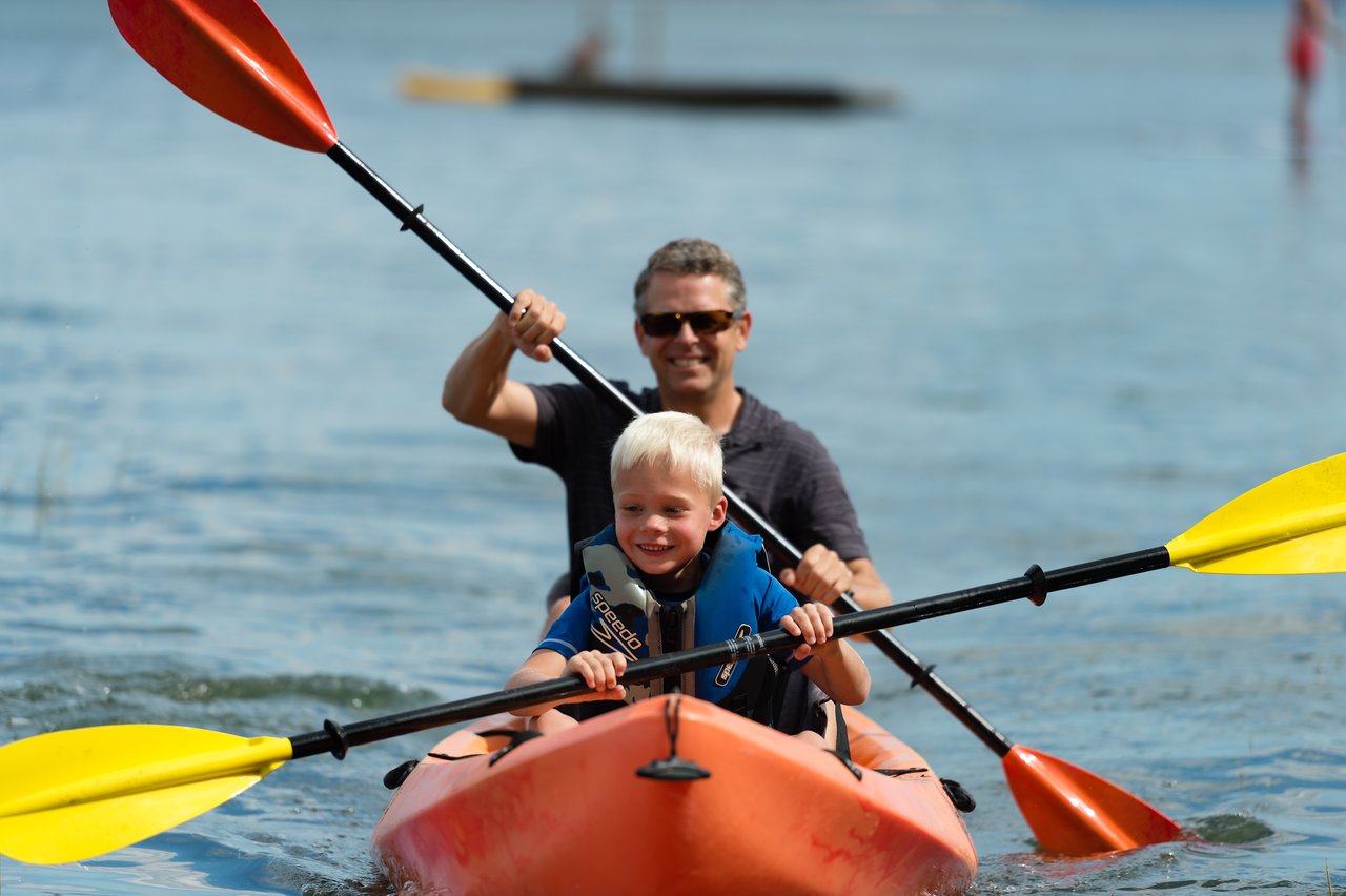 A man and a young child paddle a bright orange kayak together on calm water, both smiling.