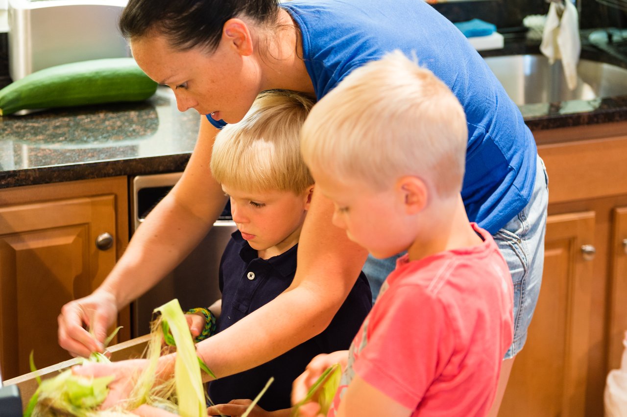 A woman and two young children husk corn together in a kitchen, preparing food for dinner.