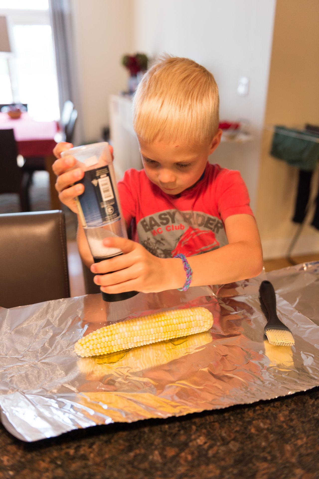 A young child in a red shirt sprinkles seasoning on an ear of corn placed on foil.