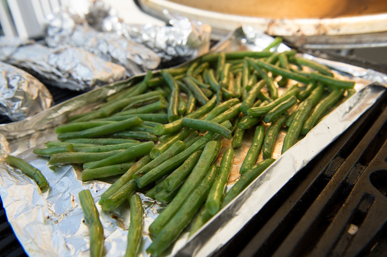 Green beans cooking on a foil-lined tray on a grill, with wrapped food in the background.