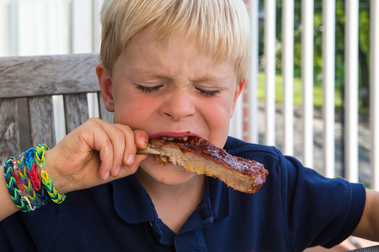 A young child in a navy shirt bites into a barbecued rib while sitting outside at a wooden table.