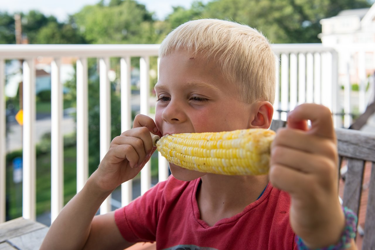 A child in a red shirt eats corn on the cob while sitting at an outdoor table.