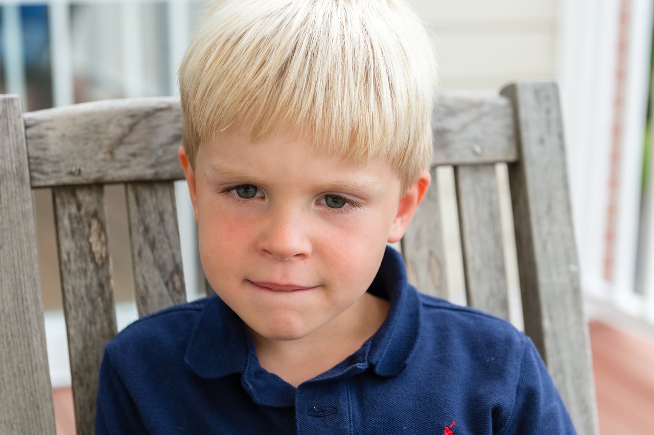 A young boy in a navy blue shirt sits on a wooden chair, biting his lip and looking ahead.