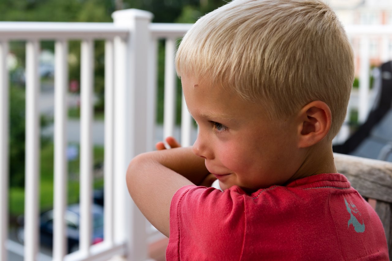 A young child in a red shirt sits on a porch, looking to the side with a slight smile.