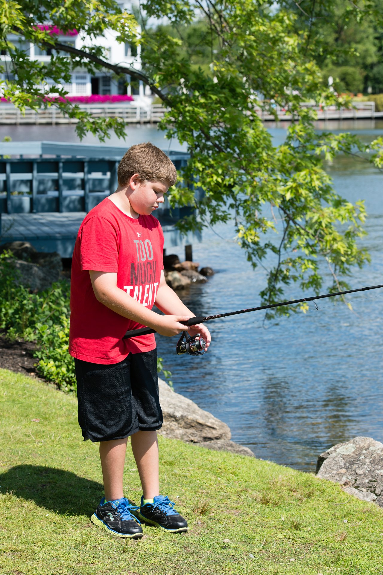 A boy in a red shirt stands by the water, holding a fishing rod and focusing on his line.