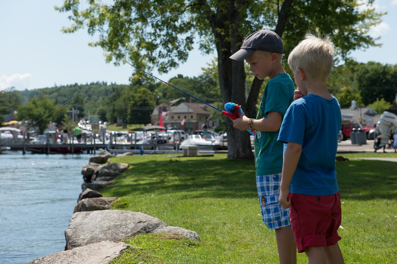 Two young boys stand near the water, one holding a fishing rod while the other watches closely.