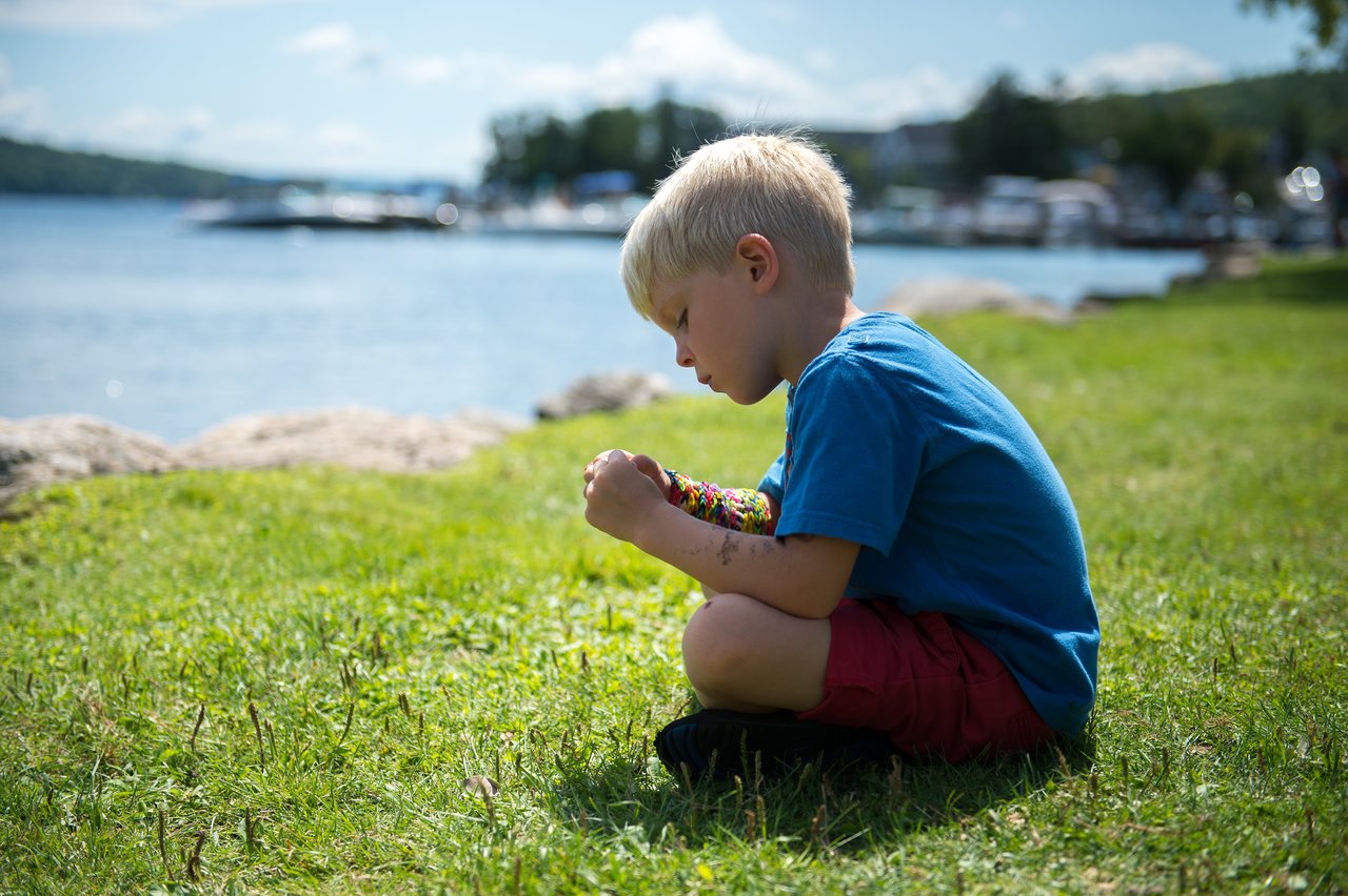 A young child sits on the grass near the water, focused on something in their hands.