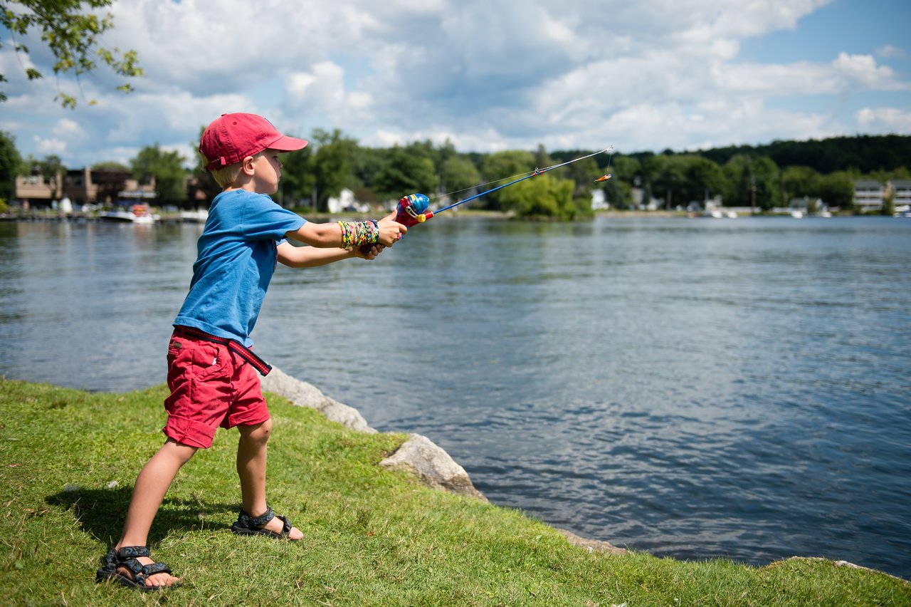 A child wearing a red cap and blue shirt casts a fishing line into the water from the grassy shore.