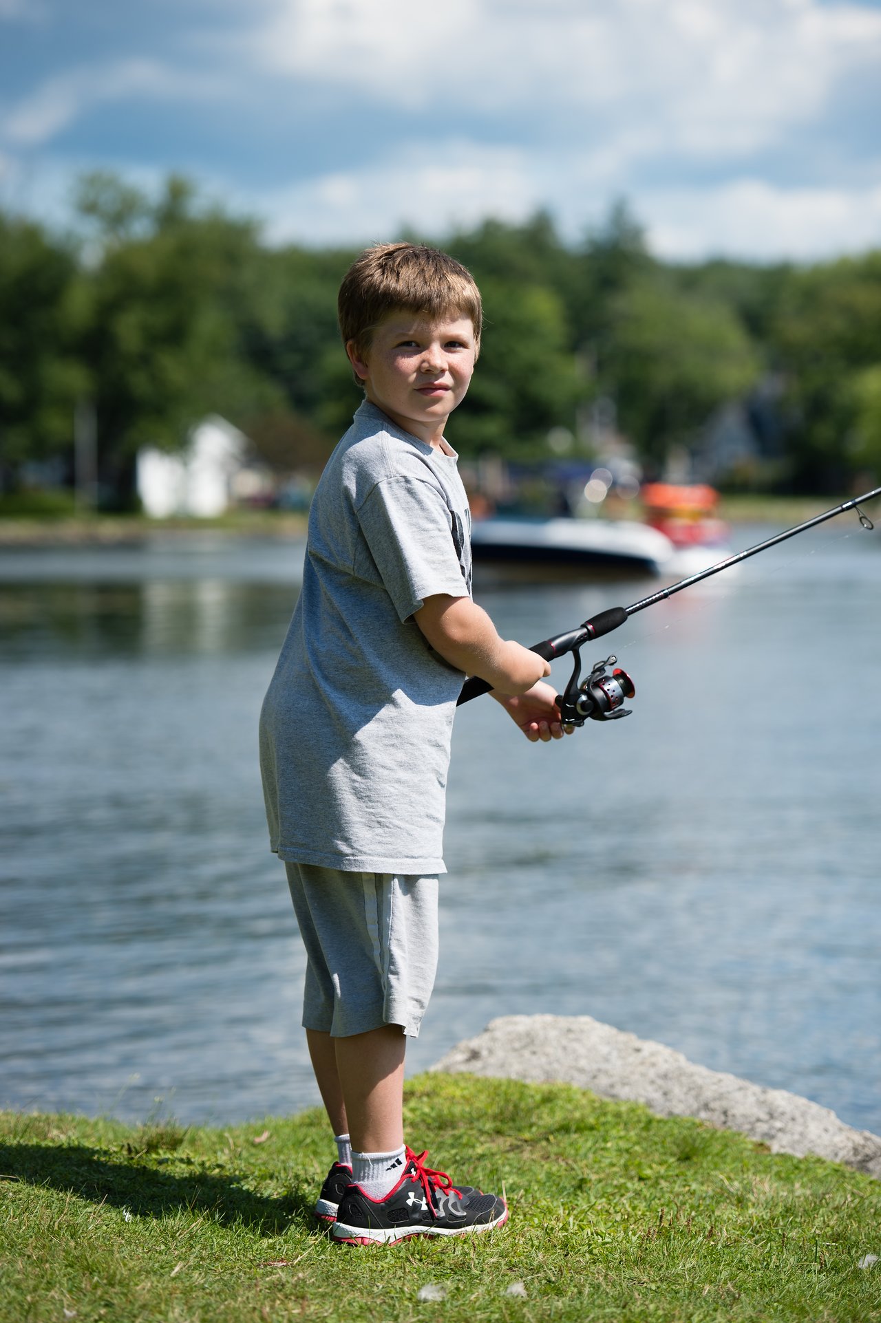 A young boy stands near the water, holding a fishing rod and looking toward the camera.