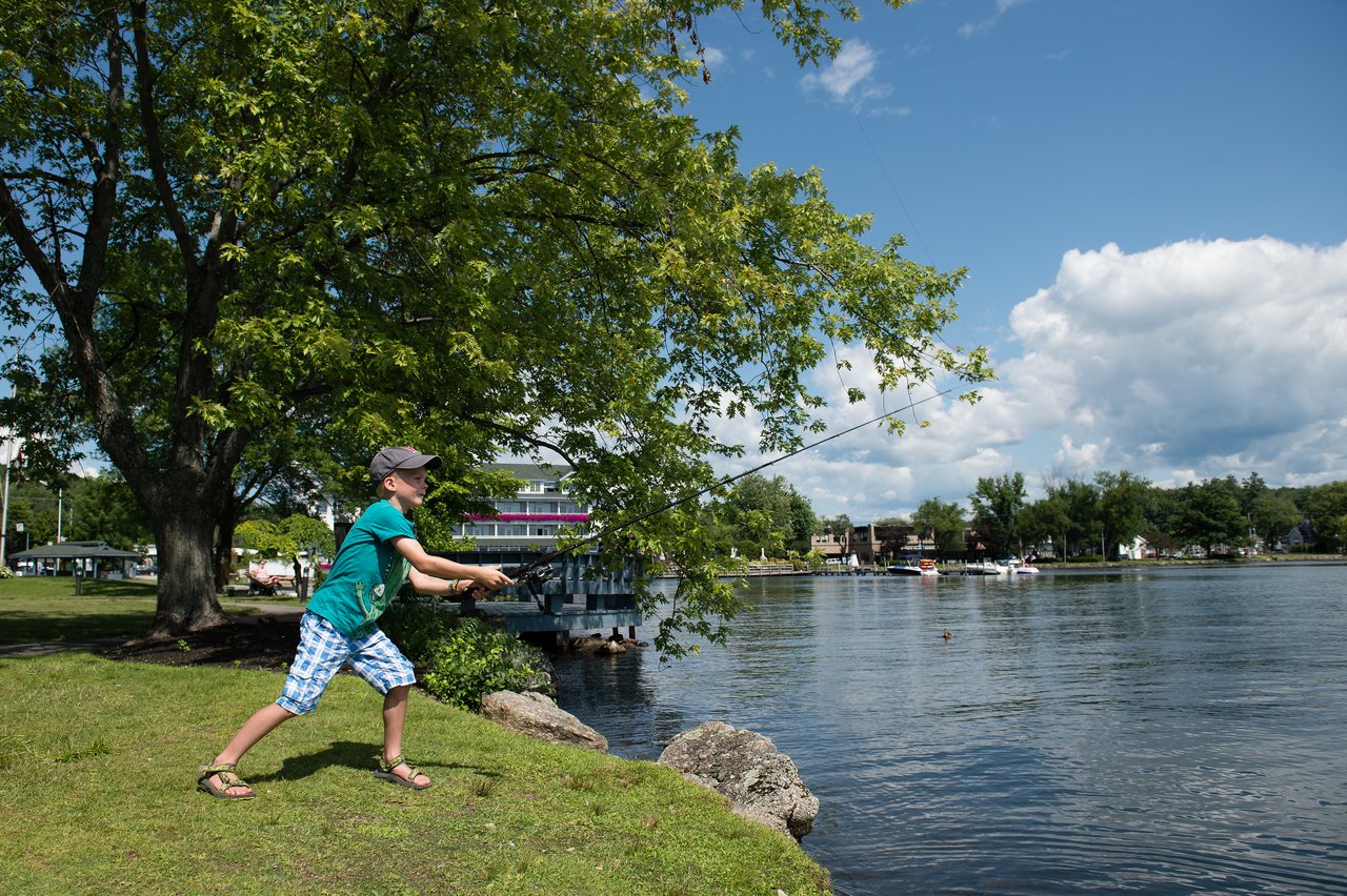 A child wearing a cap and sandals is casting a fishing line into a calm lake.