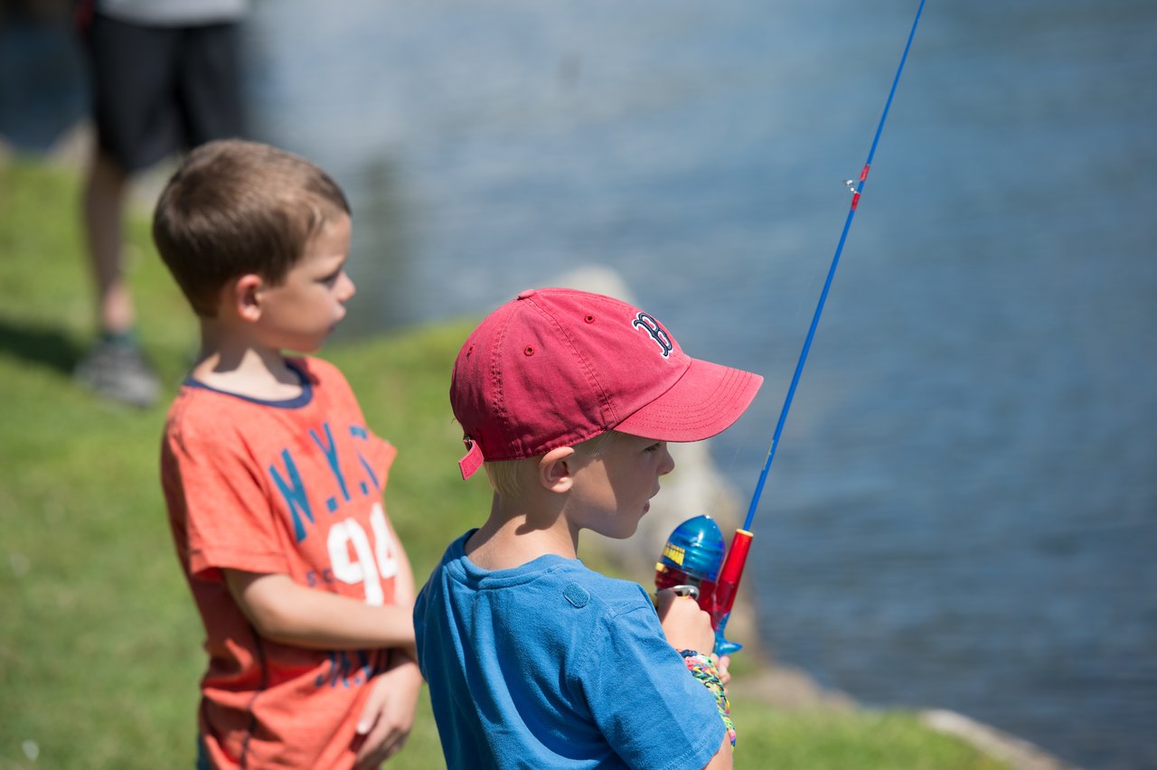 A young child in a red cap holds a fishing rod near the water, while another child watches nearby.