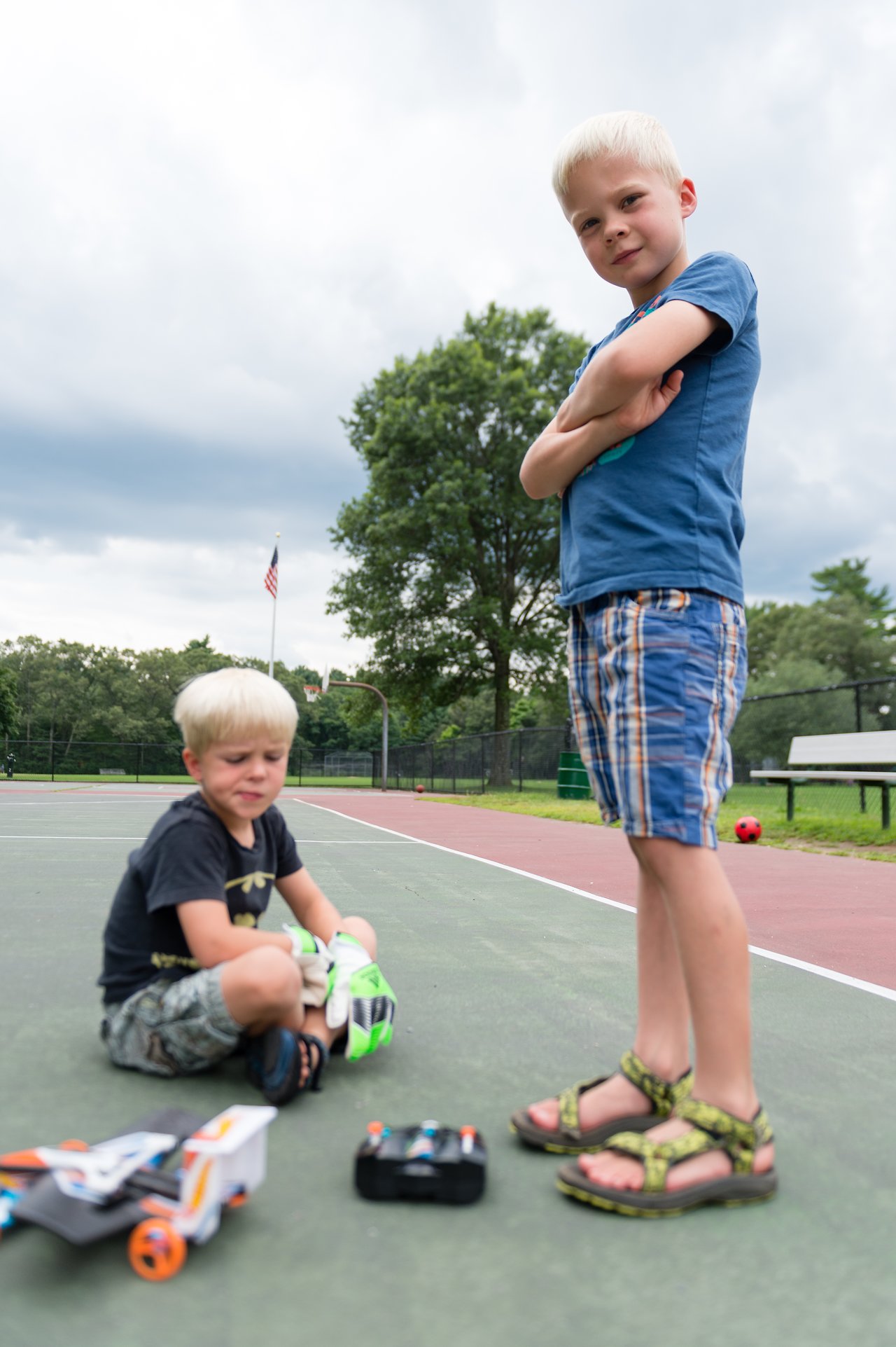 Two children on a basketball court with toy cars and a remote control, one sitting and the other standing.