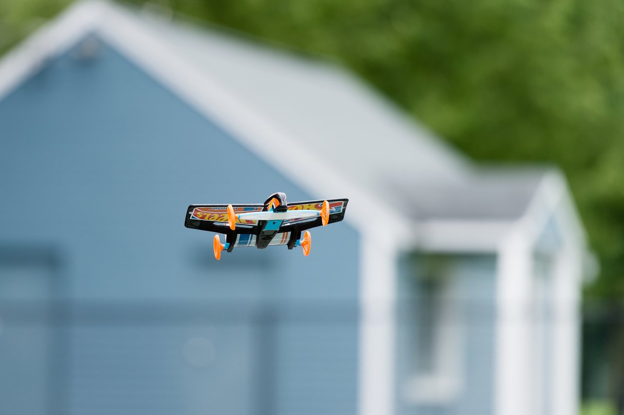 A small remote-controlled plane with orange wheels is flying in front of a blue building.