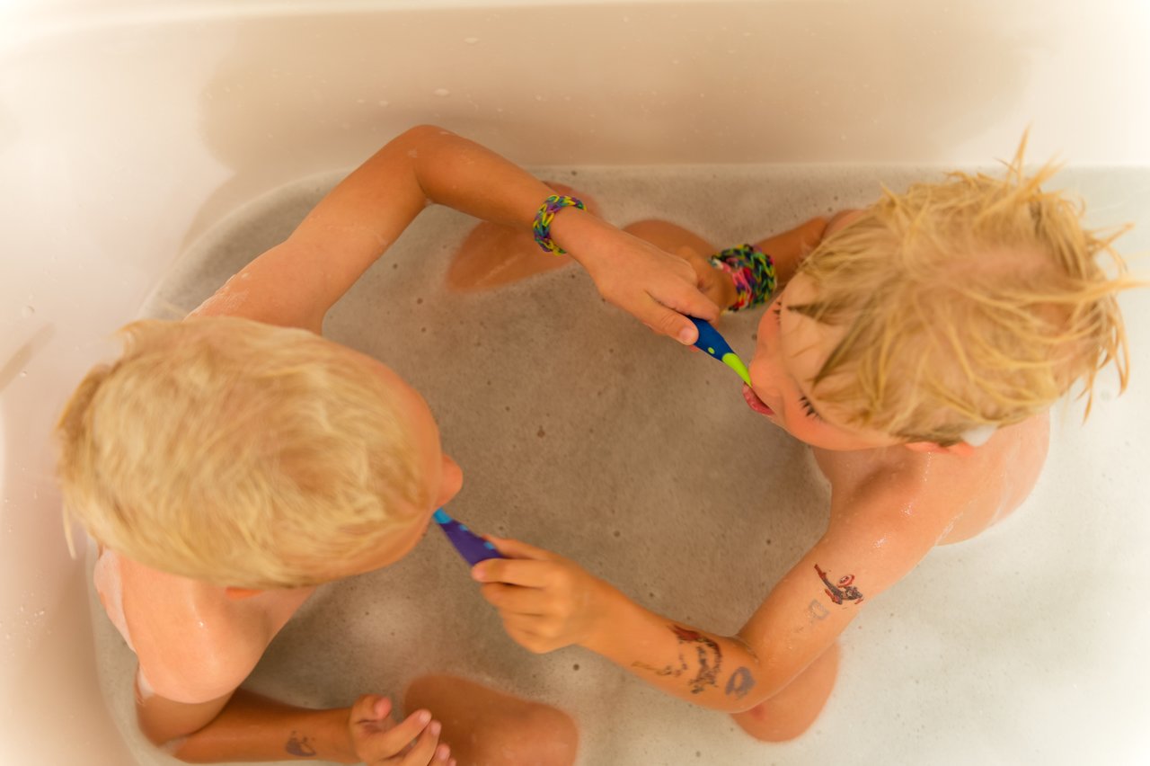 Two children in a bathtub brush each other's teeth with colorful toothbrushes.