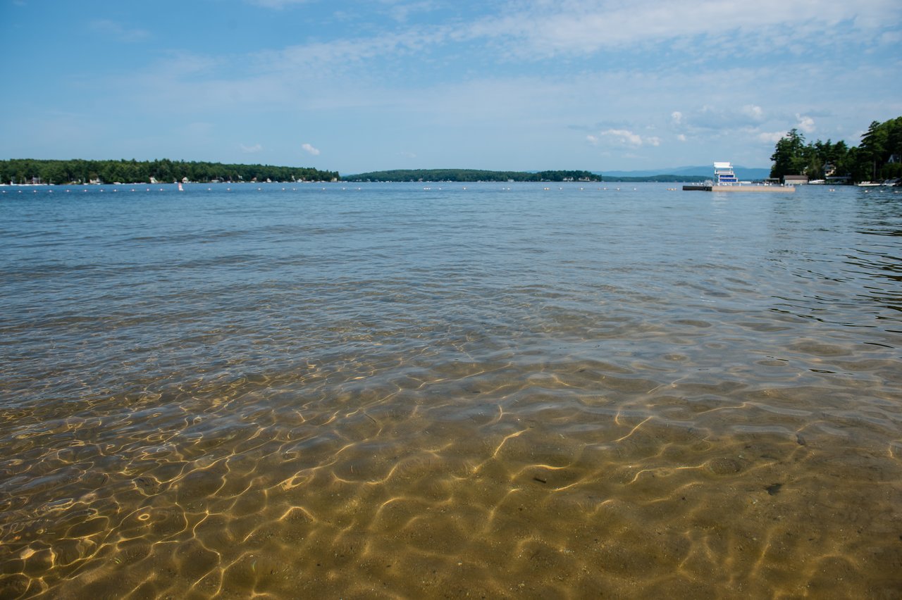Shallow, clear water at the edge of Lake Winnipesaukee with a floating dock in the distance.