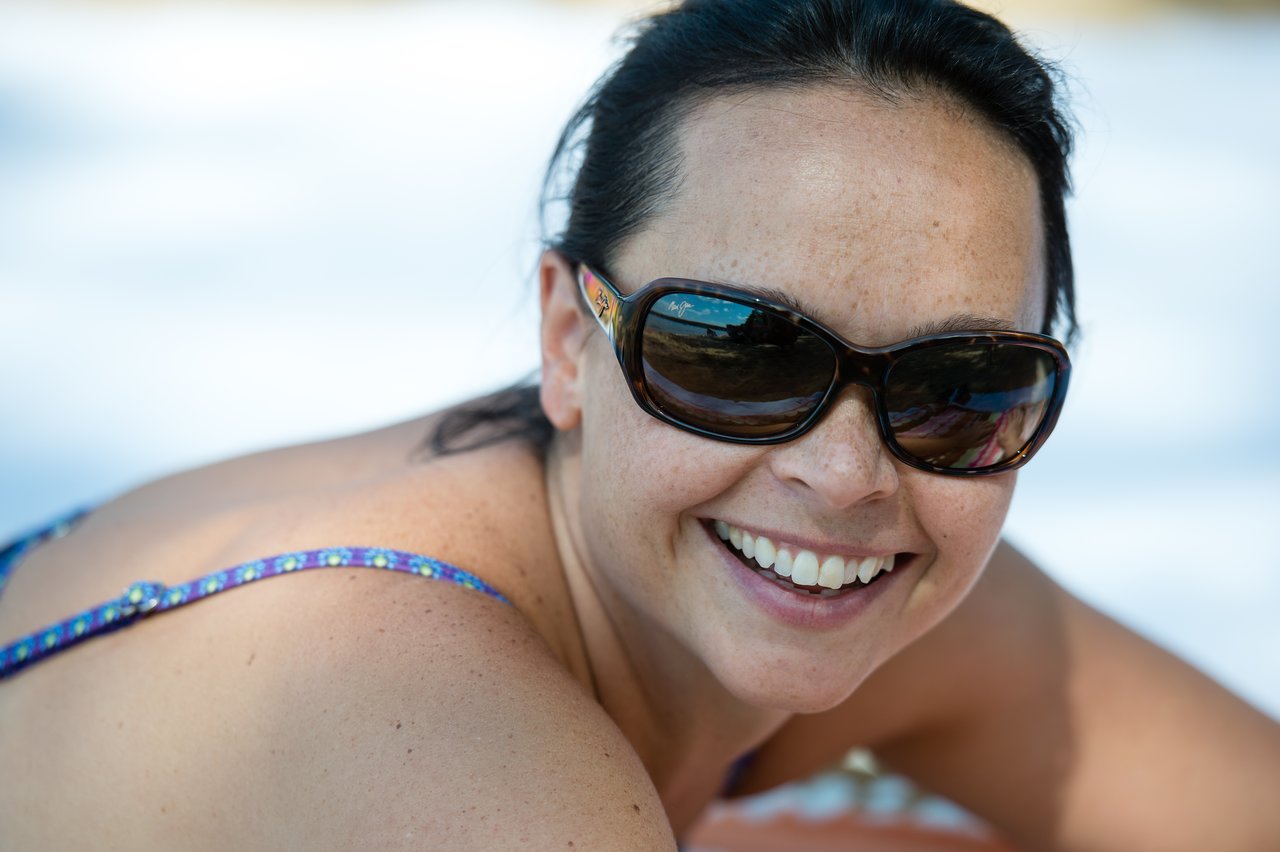 A woman wearing sunglasses and a swimsuit smiles while lying on the ground outdoors.