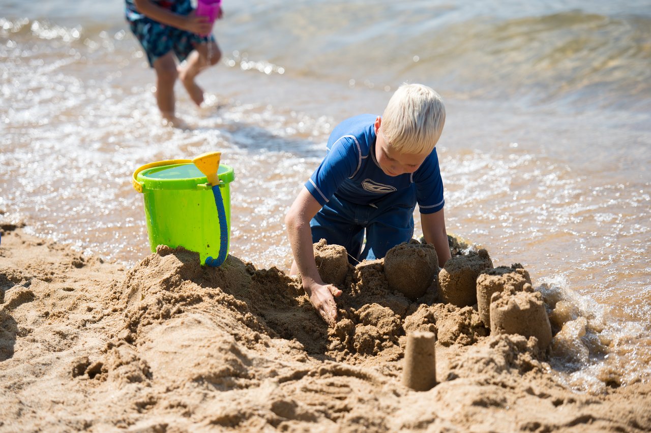 A child in a blue swimsuit builds a sandcastle on the beach near the water, with a bucket nearby.