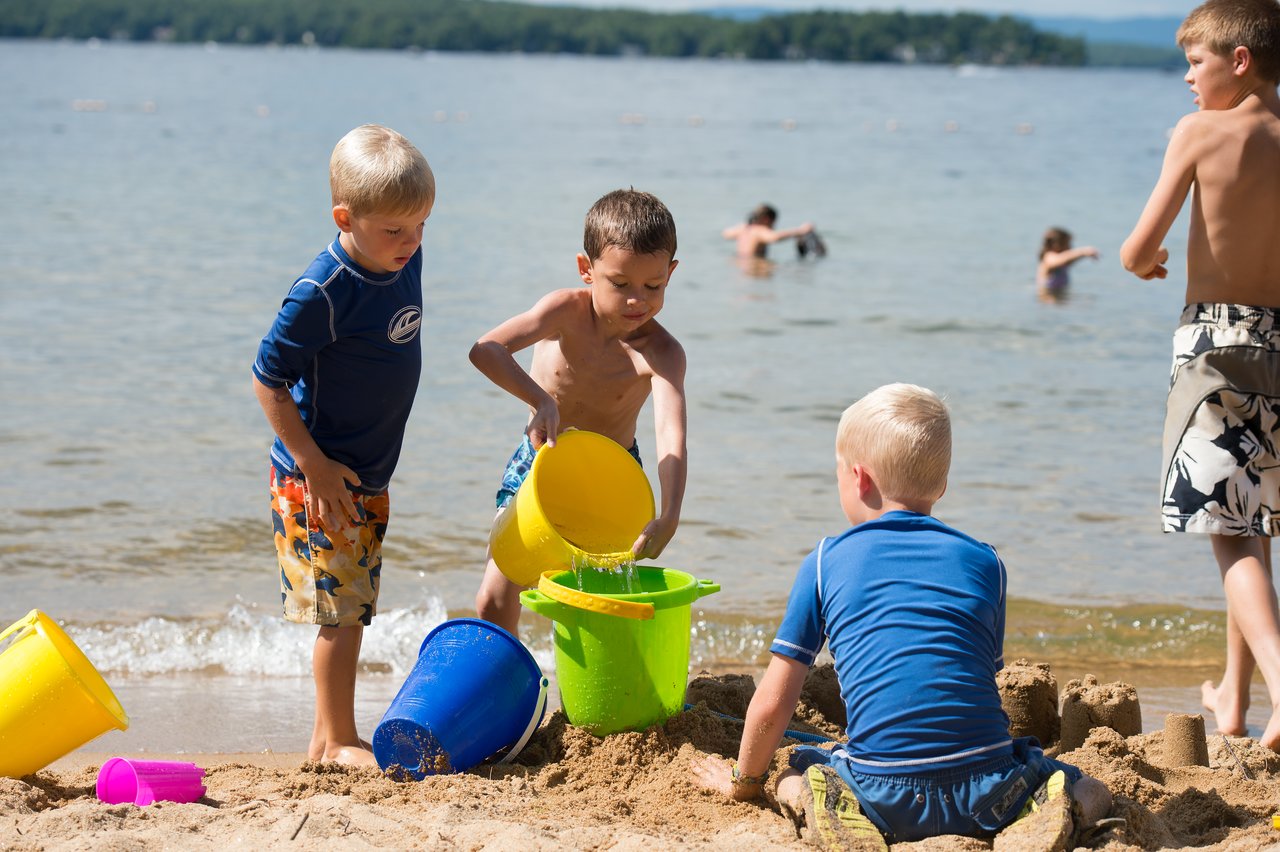 Children play in the sand by the lake, using buckets to build sandcastles and pour water.