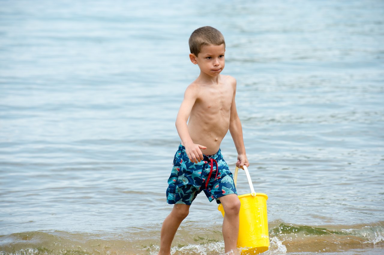 A young boy walks through shallow water carrying a yellow bucket at Lake Winnipesaukee.