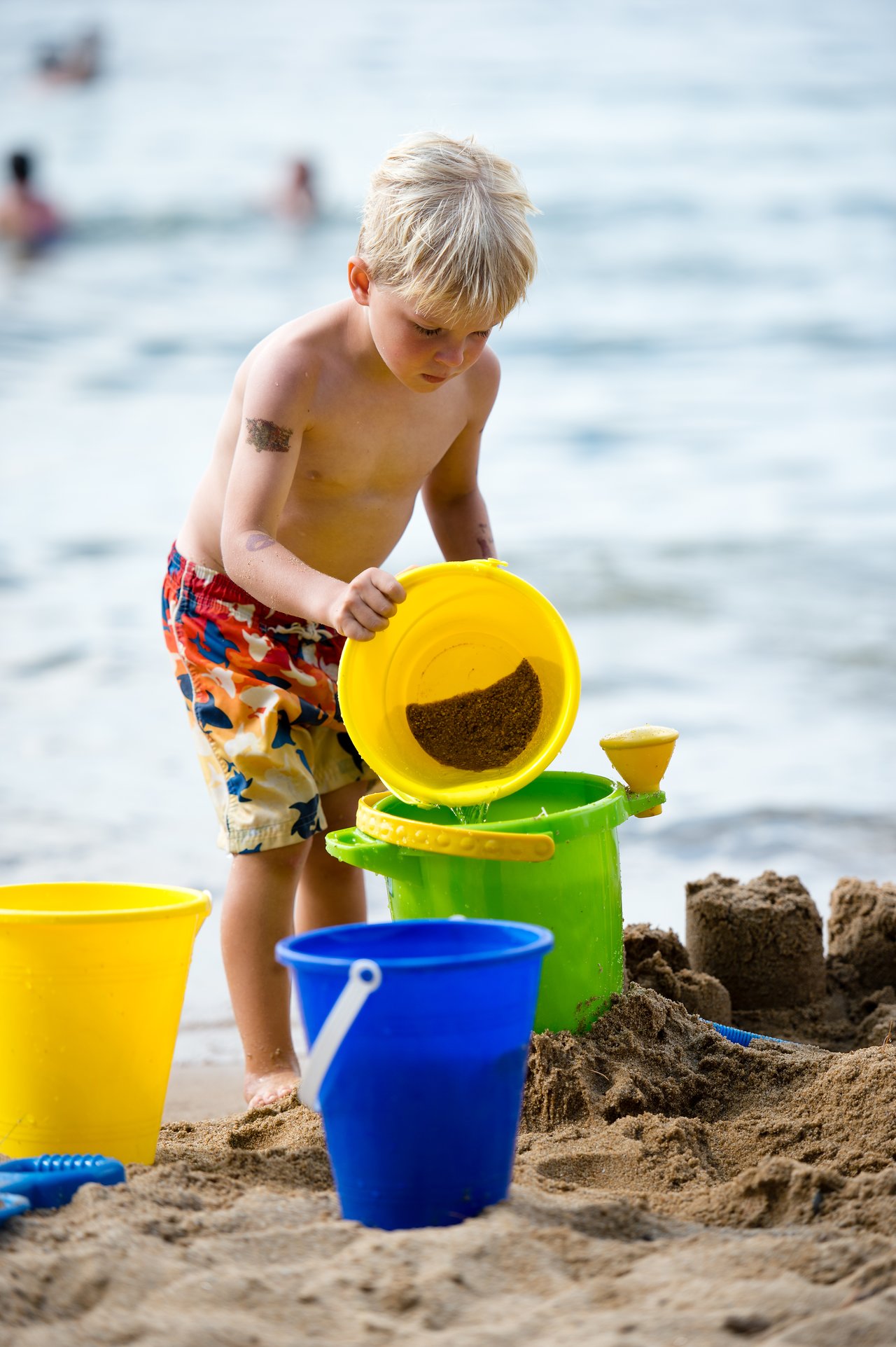 A young child pours sand from a yellow bucket while playing on the beach near the water.