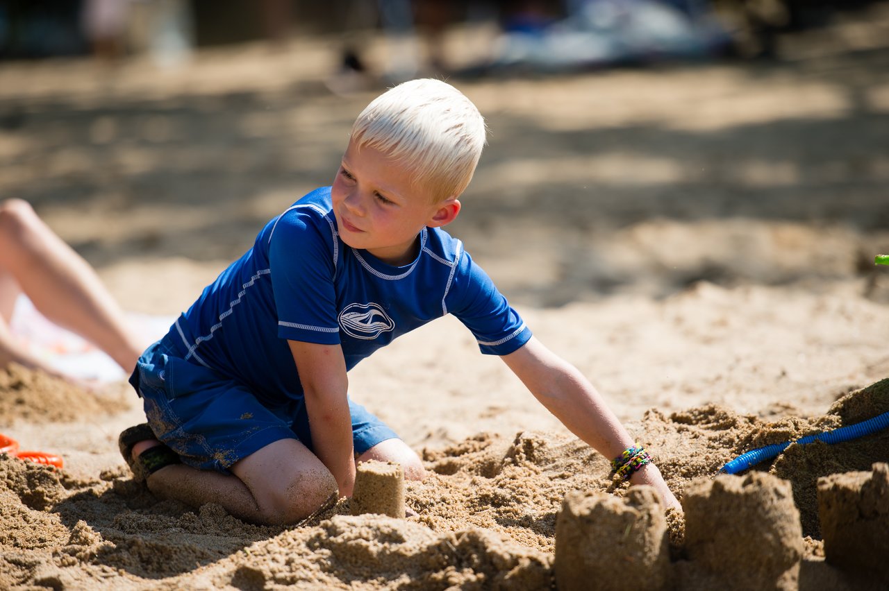 A child in a blue shirt builds a sandcastle on the beach, using their hands to shape the sand.