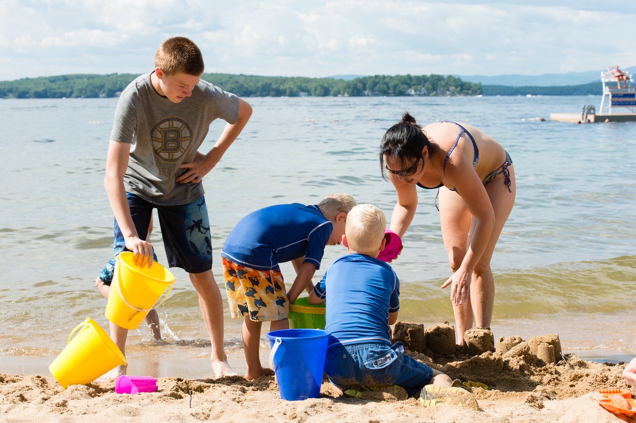 A woman and three children build a sandcastle on the beach, using buckets and shovels near the water.