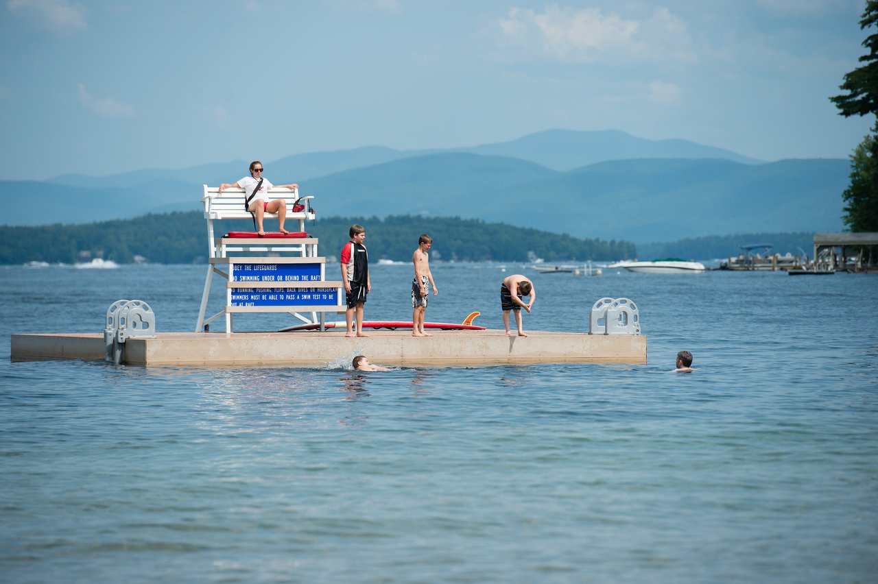 A lifeguard watches from a tall chair as children stand, swim, and prepare to jump off a floating dock.