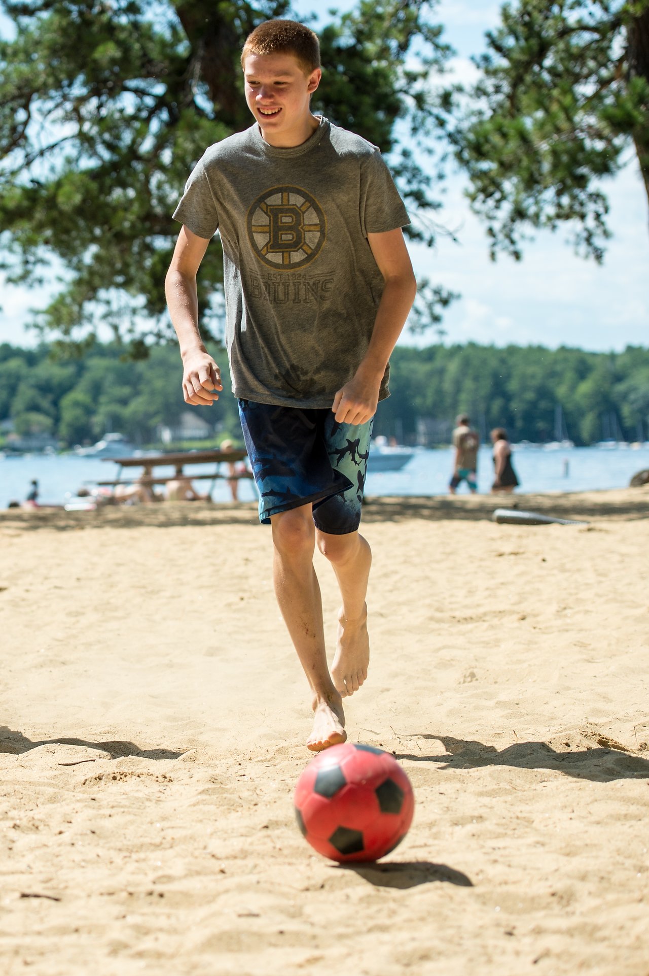 A barefoot boy plays soccer on a sandy beach, kicking a red and black ball near the lake.