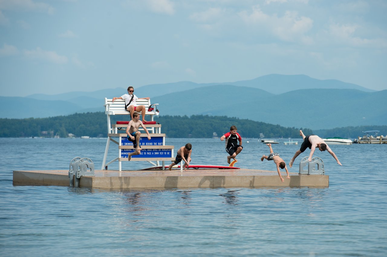 People on a floating dock at Lake Winnipesaukee, with some jumping into the water and a lifeguard watching.