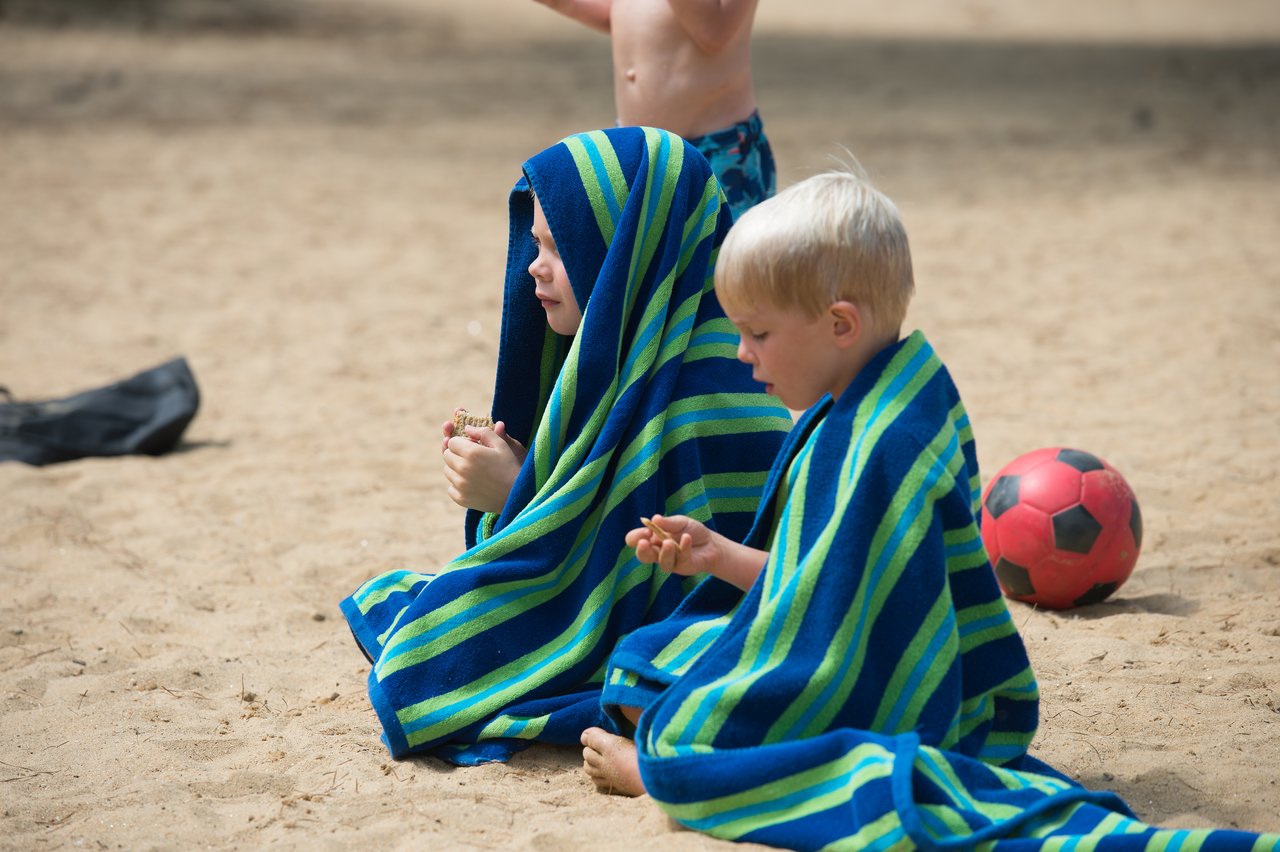 Two young children sit on the sandy beach, wrapped in striped towels, eating snacks.